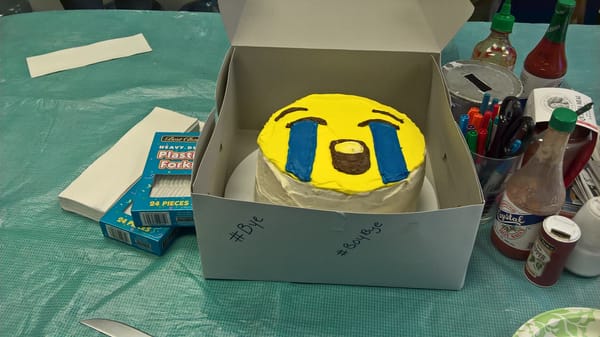 A beautiful cake that looks like the bawling emoji, on a table with plastic forks, crystal sauce, and various other clutter